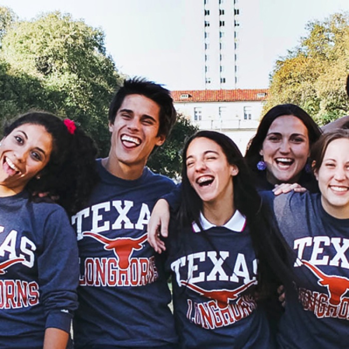 A group of students smile and link arms wearing matching Texas t-shirts in front of the tower.