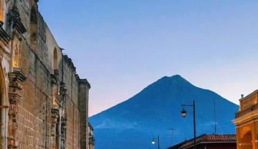 A perspective view of an old stone street and historic buildings lit up in the foreground framing the distant Agua volcano in Antigua, Guatemala during the blue hour of the evening.
